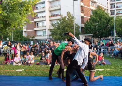 Quatre artistes posent pour un selfie sur un tapis bleu en plein air, avec un public nombreux assis et des immeubles d'habitation en arrière-plan.