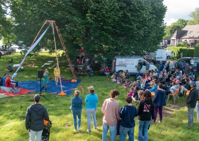 Un artiste de cirque en plein air se tient sous un dispositif aérien tandis qu'un public assis l'observe. Des gens sont rassemblés sur l'herbe, et une camionnette est garée à proximité, sous de grands arbres.