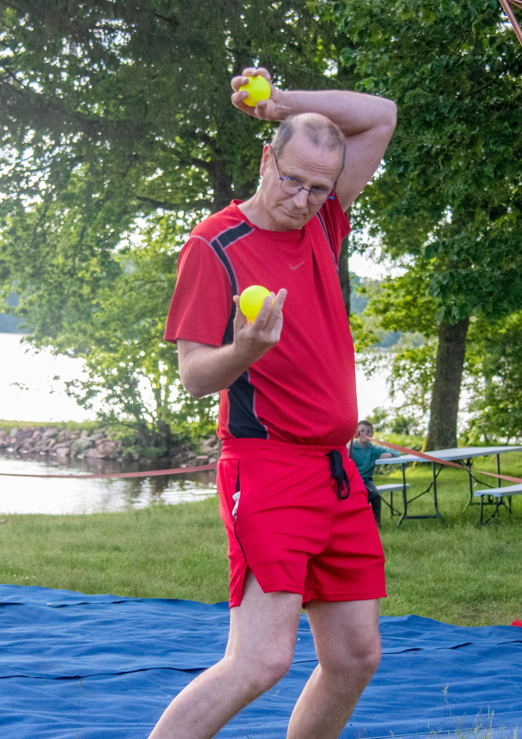 Un homme vêtu d'une tenue de sport rouge jongle avec des balles jaunes à l'extérieur, sur un tapis bleu, avec des arbres, de l'herbe et de l'eau en arrière-plan.